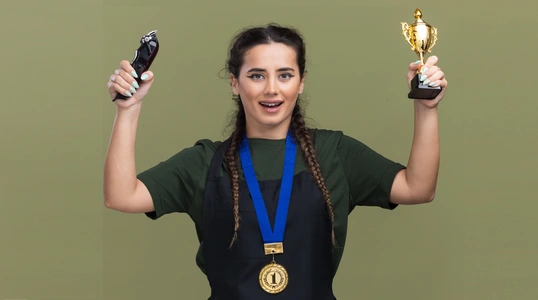 smiling young female barber uniform medal raising winner cup with hair clippers isolated olive green wall